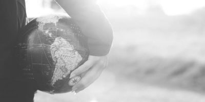Person standing looking at the sun set holding a globe in their right hand against their hip